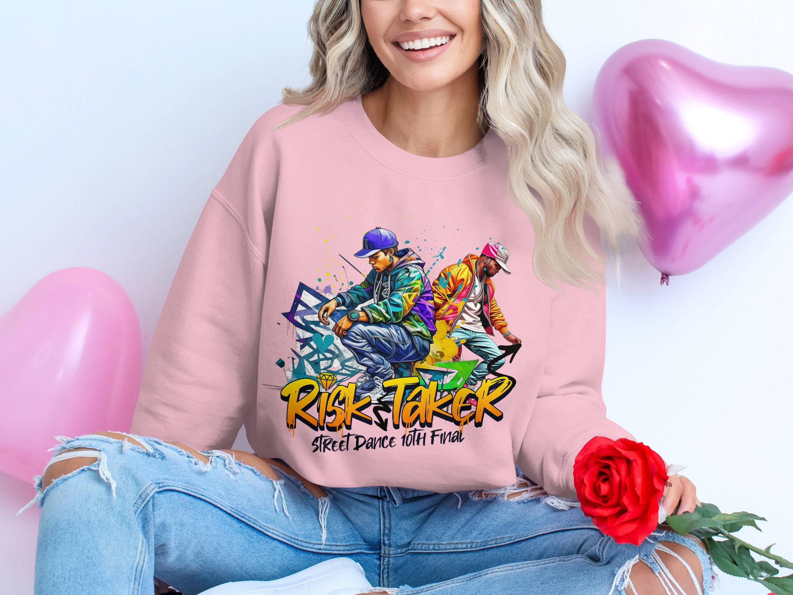 A person in a Garment Graphics pink sweatshirt with the print, Risk Taker Street Dance 10th Final, holds a red rose, smiling in ripped jeans amid pink heart-shaped balloons, celebrating the street dance finals.