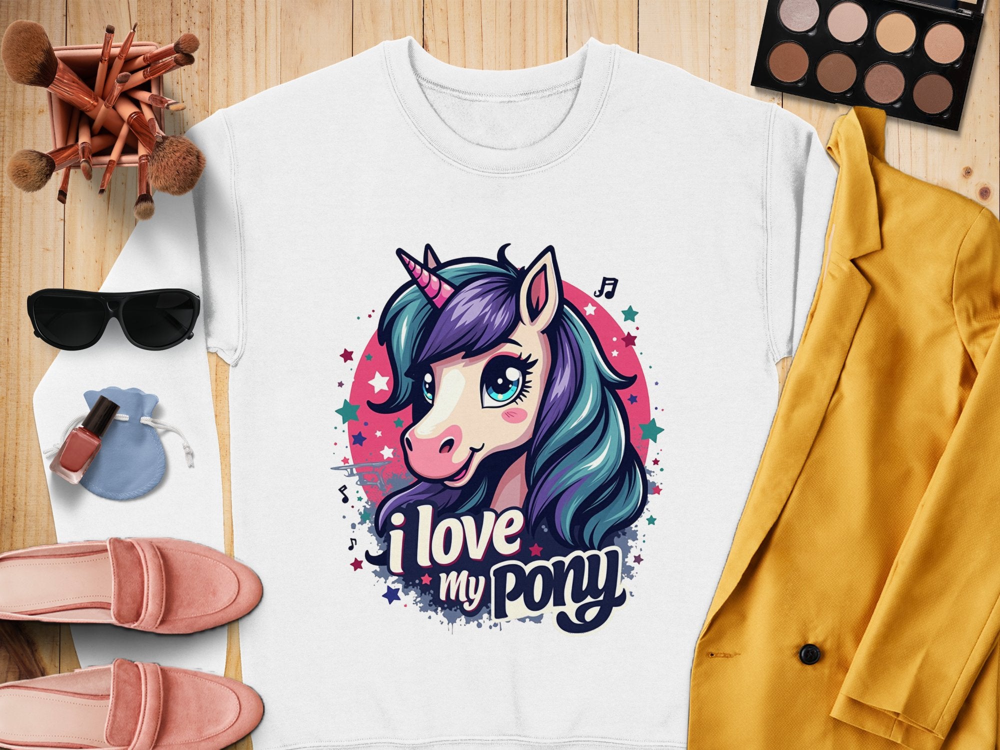 Flat lay of a Garment Graphics white graphic sweatshirt with a ponyunicorn design and I love my pony text, surrounded by sunglasses, makeup brushes, a palette, pink loafers, blue drawstring pouch, nail polish, and yellow jacket on wooden surface.