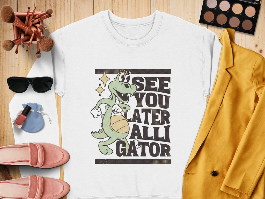 A fun Garment Graphics white sweatshirt features an alligator graphic with See You Later Alligator text, styled elegantly with sunglasses, makeup brushes, a palette, lipstick, pink shoes, and a mustard yellow blazer on a wooden surface.
