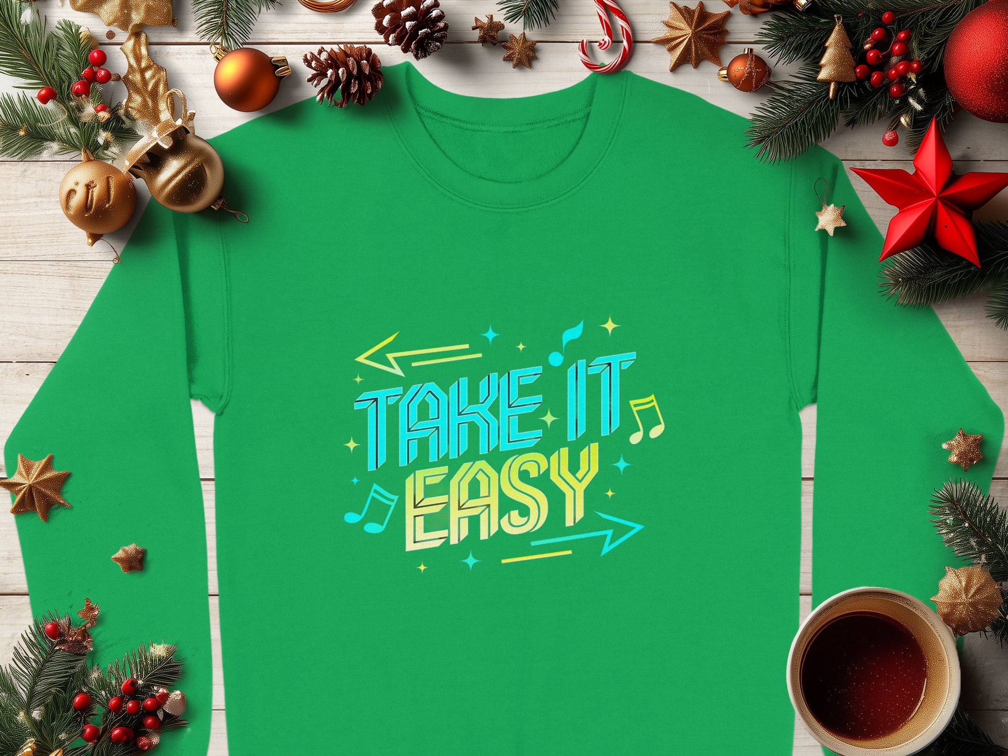 Garment Graphics green sweatshirt features a vibrant Take It Easy graphic with musical notes and arrows. Displayed on a wooden surface with pine branches, ornaments, and a steaming hot beverage, it offers unisex sizing for a relaxed fit.
