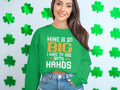 In front of a shamrock-decorated wall, a woman with long brown hair joyfully wears Garment Graphics unique green sweatshirt adorned with white and orange text: Mine Is So Big I Have To Use Both Hands.