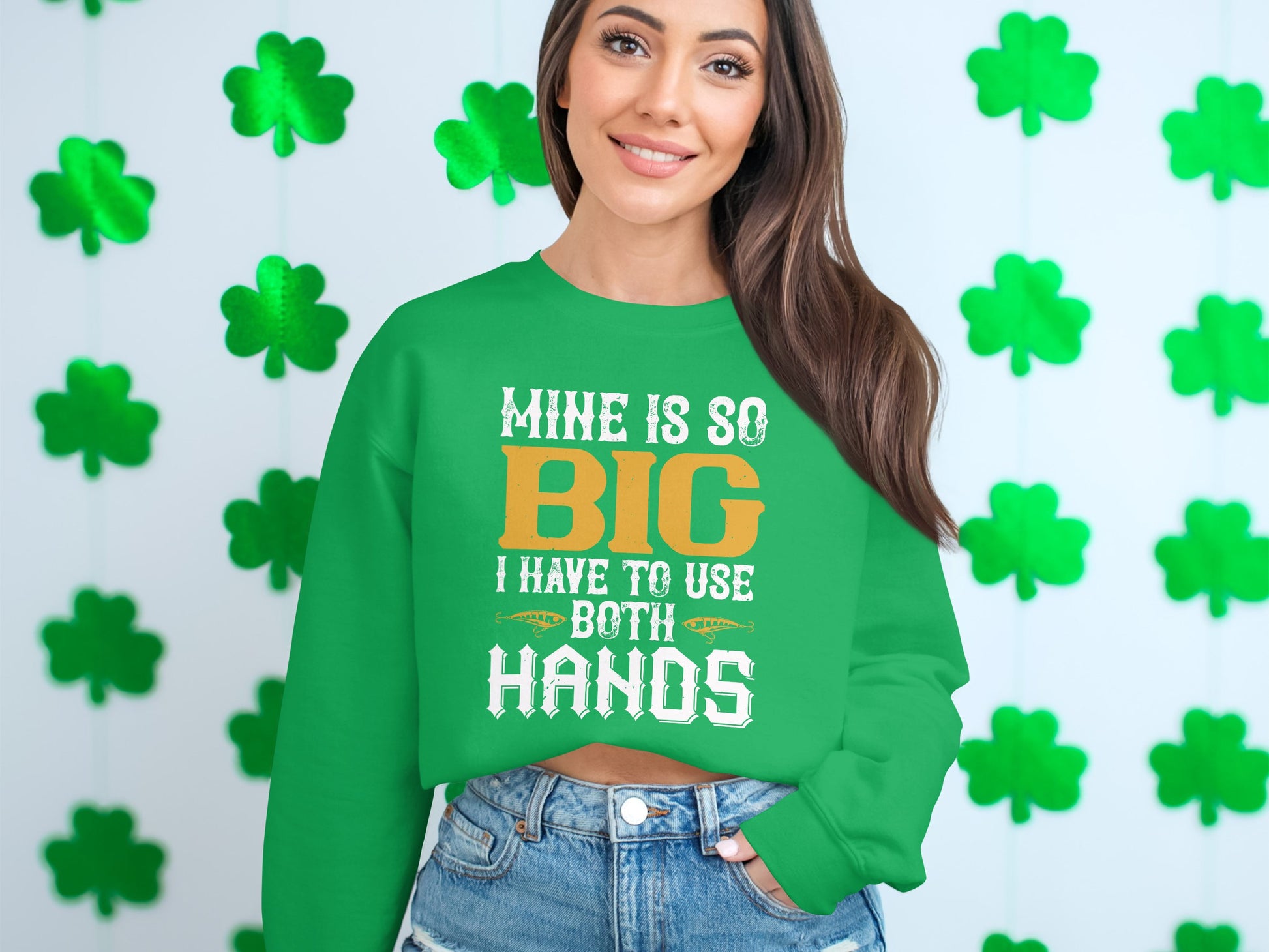 In front of a shamrock-decorated wall, a woman with long brown hair joyfully wears Garment Graphics unique green sweatshirt adorned with white and orange text: Mine Is So Big I Have To Use Both Hands.