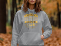 A person in a light gray Garment Graphics golf hoodie with a humorous statement: If 45s got you climbing the tree, TRICK OR TREAT a camper in. They dont hibernate, stands hands in pockets against gently swaying blurred fall foliage.