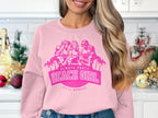In a festive kitchen filled with Christmas decorations, someone wears a pink sweatshirt from Garment Graphics featuring the Always Party Beach theme and an illustration of three girls. Small pine trees and ornaments complete the cheerful holiday scene.