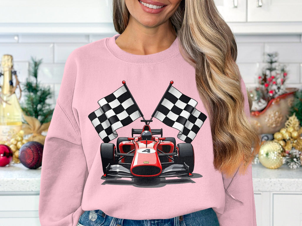 In a festive kitchen with a small tree and ornaments, a woman wears a vibrant pink sweatshirt featuring a red racing car and checkered flags, perfect for motorsport fans. The sweatshirt is by Garment Graphics.