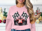 In a festive kitchen with a small tree and ornaments, a woman wears a vibrant pink sweatshirt featuring a red racing car and checkered flags, perfect for motorsport fans. The sweatshirt is by Garment Graphics.