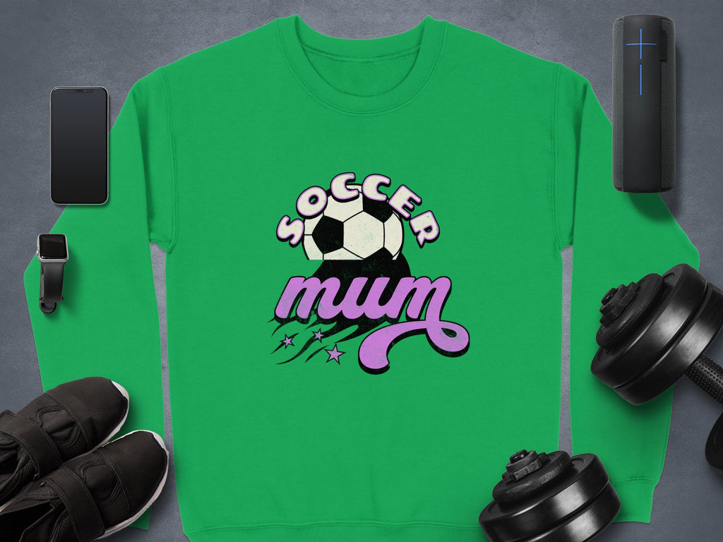 A vintage-style green sweatshirt by Garment Graphics features Soccer Mum, a soccer ball, and purple stars, surrounded by a smartphone, smartwatch, sneakers, wireless speaker, and dumbbells on a gray background.