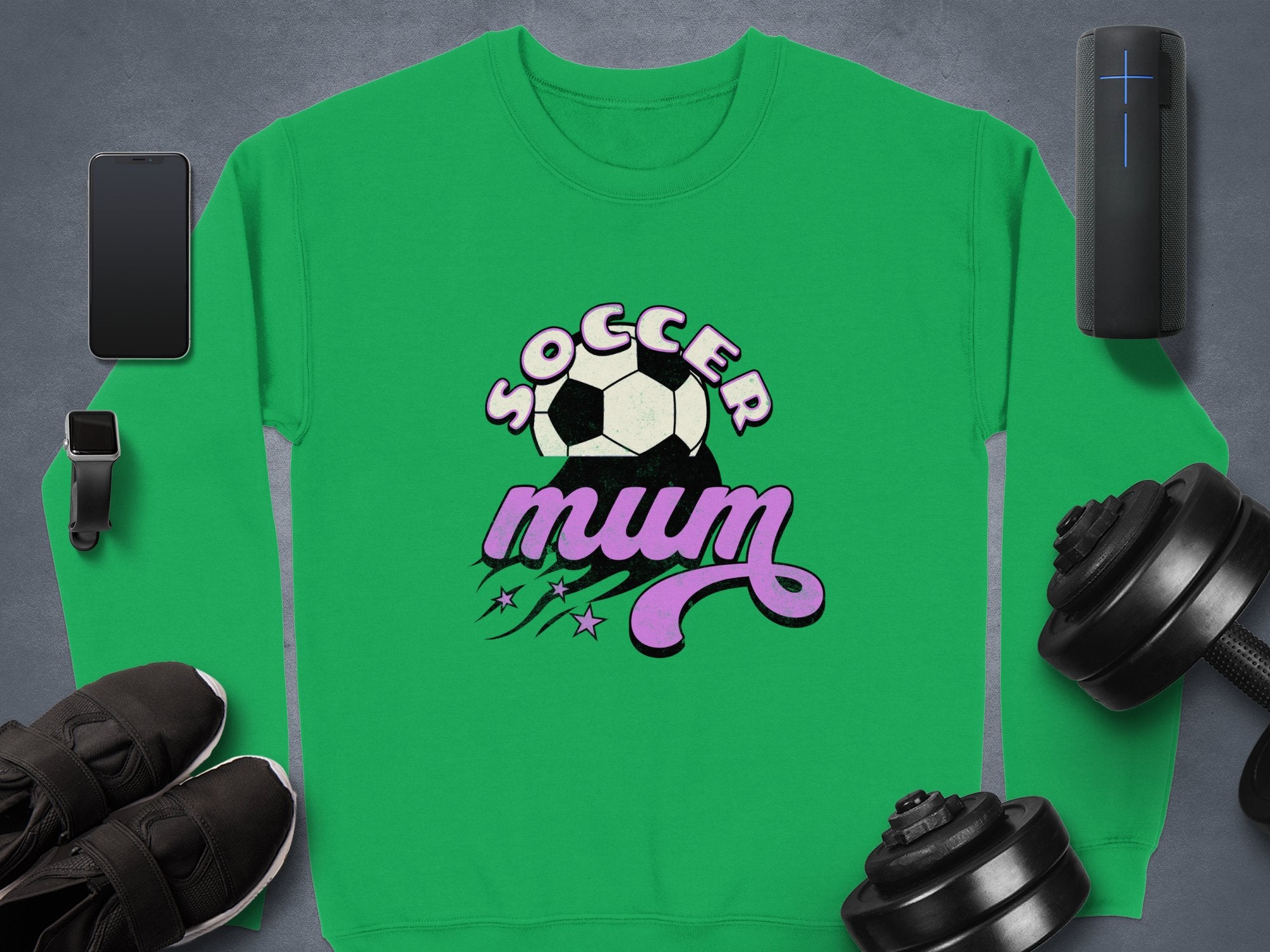 A vintage-style green sweatshirt by Garment Graphics features Soccer Mum, a soccer ball, and purple stars, surrounded by a smartphone, smartwatch, sneakers, wireless speaker, and dumbbells on a gray background.