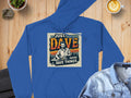 A blue graphic hoodie from Garment Graphics, featuring Just Dave Doing Dave Things in a retro font, is made of medium-heavy fabric and rests on a wooden surface beside a denim jacket, coffee, and a potted plant.