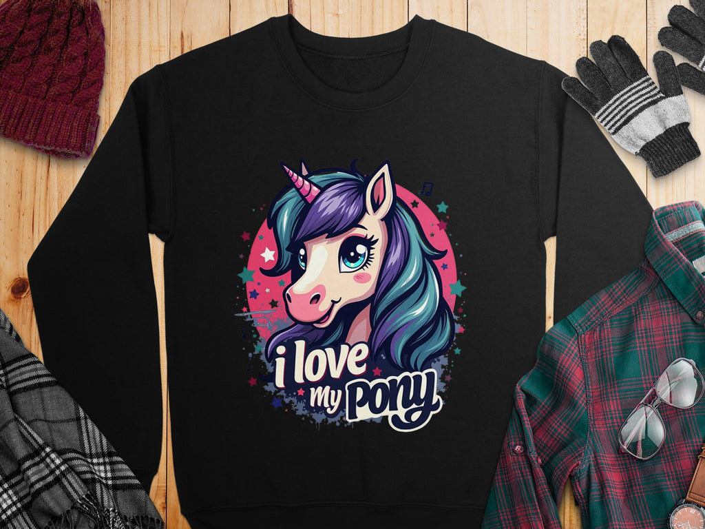Garment Graphics sweatshirt features a vibrant cartoon unicorn with a blue and purple mane, pink horn, large eyes surrounded by stars, and the text I love my pony. Its displayed on a wooden surface with a hat, gloves, scarf, and plaid shirt.