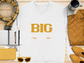 A Garment Graphics white t-shirt with BIG in yellow letters and two fish illustrations is on a wooden surface with a plaid scarf, notebook, pen, coffee cup, yellow quilted bag, sunglasses, and jacket—a bold piece within everyday essentials.