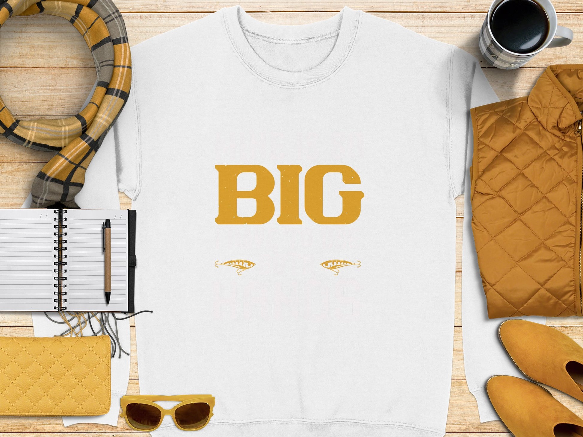 A Garment Graphics white t-shirt with BIG in yellow letters and two fish illustrations is on a wooden surface with a plaid scarf, notebook, pen, coffee cup, yellow quilted bag, sunglasses, and jacket—a bold piece within everyday essentials.