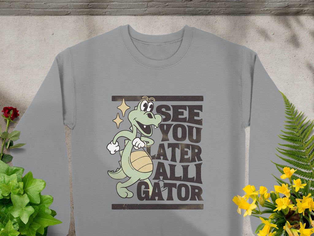 The Garment Graphics gray sweatshirt features a cartoon alligator holding a basketball with the phrase See You Later Alligator, surrounded by potted plants on concrete—ideal for fans of playful alligator designs.