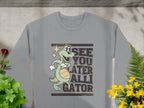 The Garment Graphics gray sweatshirt features a cartoon alligator holding a basketball with the phrase See You Later Alligator, surrounded by potted plants on concrete—ideal for fans of playful alligator designs.