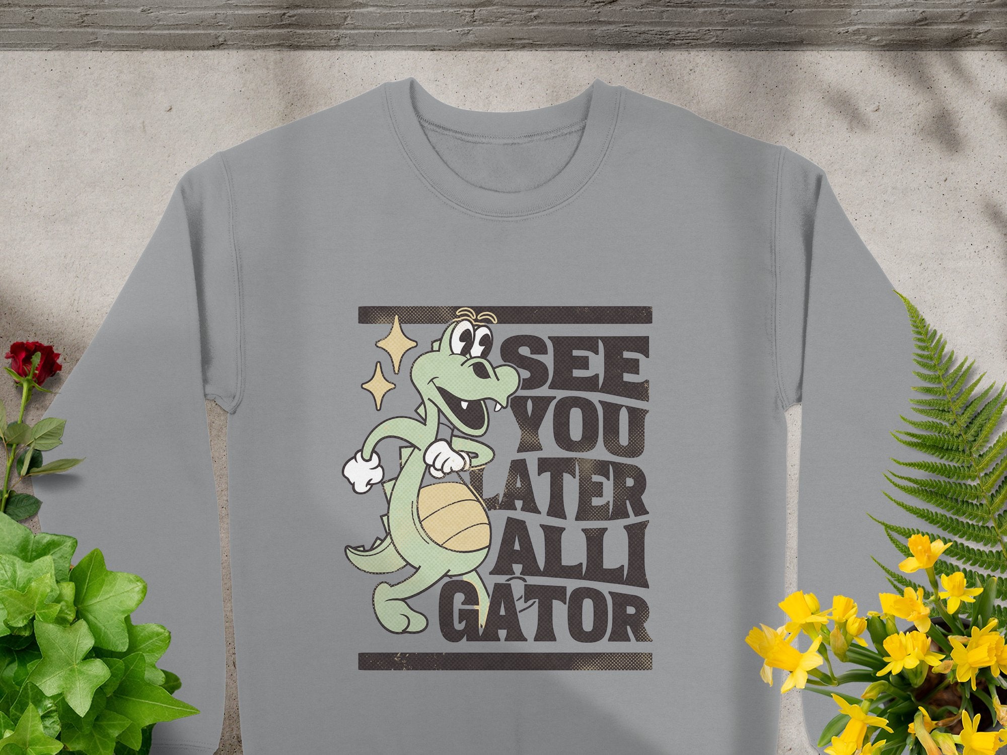 The Garment Graphics gray sweatshirt features a cartoon alligator holding a basketball with the phrase See You Later Alligator, surrounded by potted plants on concrete—ideal for fans of playful alligator designs.