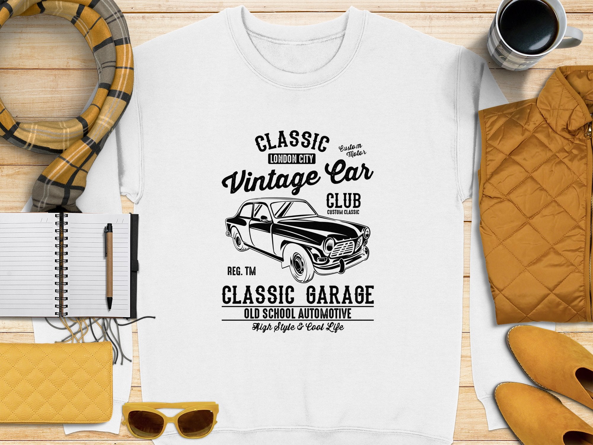 A flat lay showcases Garment Graphics white T-shirt, featuring a vintage car design and Classic London City Vintage Car Club, Classic Garage text. The scene includes a yellow purse, sunglasses, notebook, pen, plaid scarf, and steaming coffee.