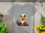 Garment Graphics presents a gray eagle T-shirt with a cartoon eagle showing a thumbs-up and green eyes. Ideal for animal lovers, its showcased on a concrete-like surface surrounded by green and yellow foliage, with a red flower in the top left corner.