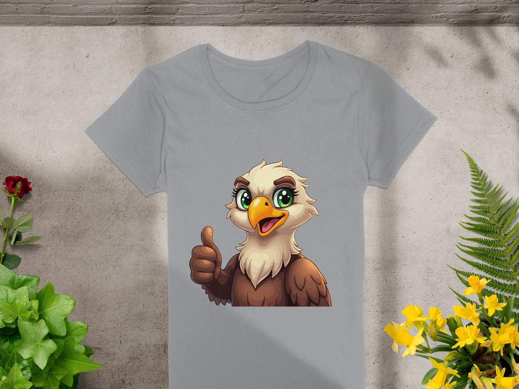Garment Graphics presents a gray eagle T-shirt with a cartoon eagle showing a thumbs-up and green eyes. Ideal for animal lovers, its showcased on a concrete-like surface surrounded by green and yellow foliage, with a red flower in the top left corner.