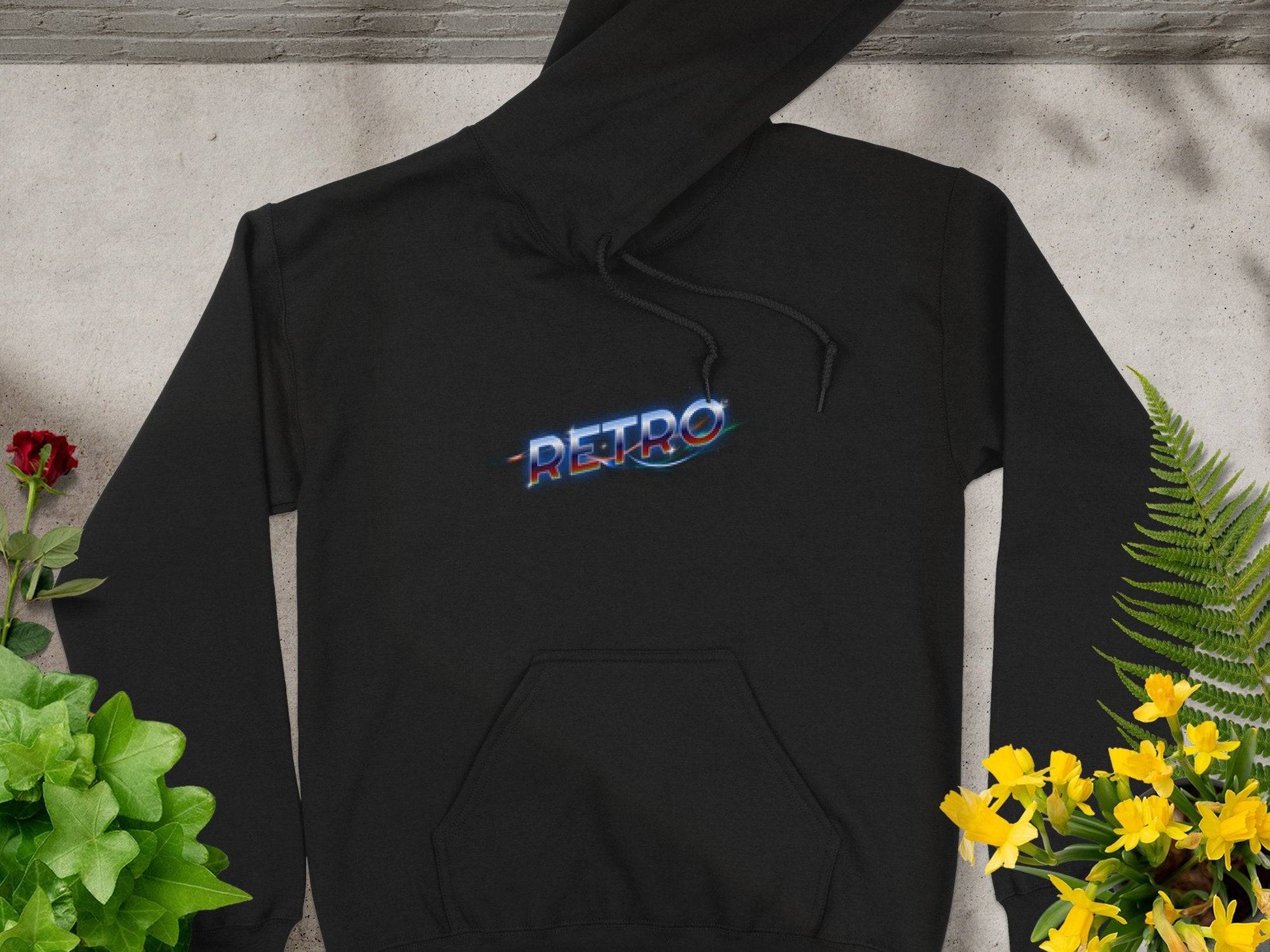 The Garment Graphics black hoodie features RETRO in a neon-style front. Laid flat on concrete, it is surrounded by vibrant green plants, red roses, and yellow flowers, capturing a trendy modern style.