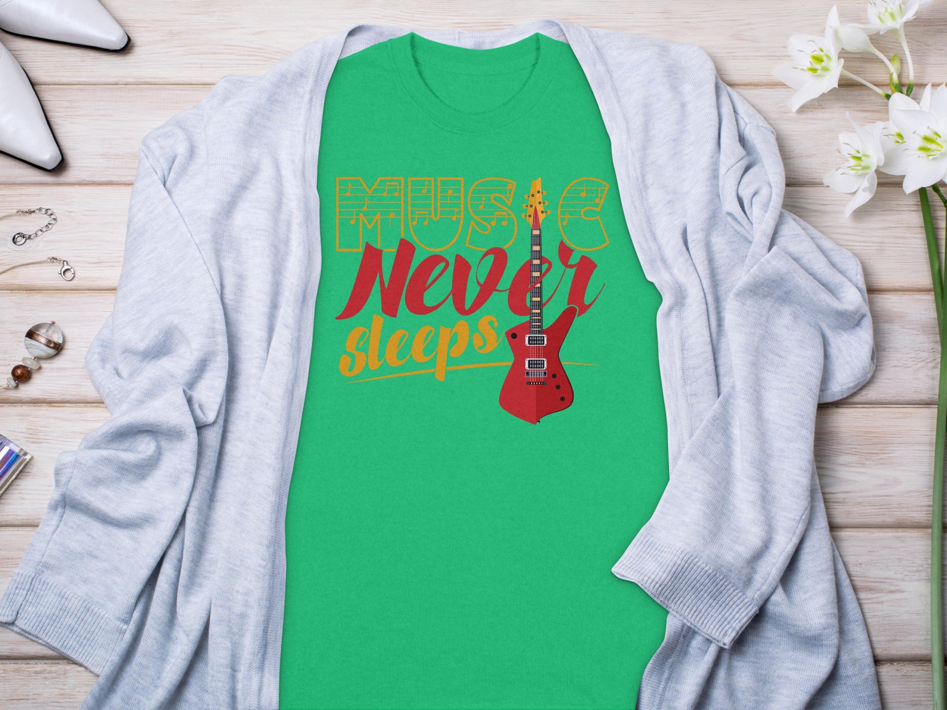 A green graphic T-shirt by Garment Graphics, crafted from 100% cotton with Music Never Sleeps and an electric guitar design, is stylishly arranged under a gray cardigan on a light wood surface, complemented by white flowers, high heels, and delicate jewelry.