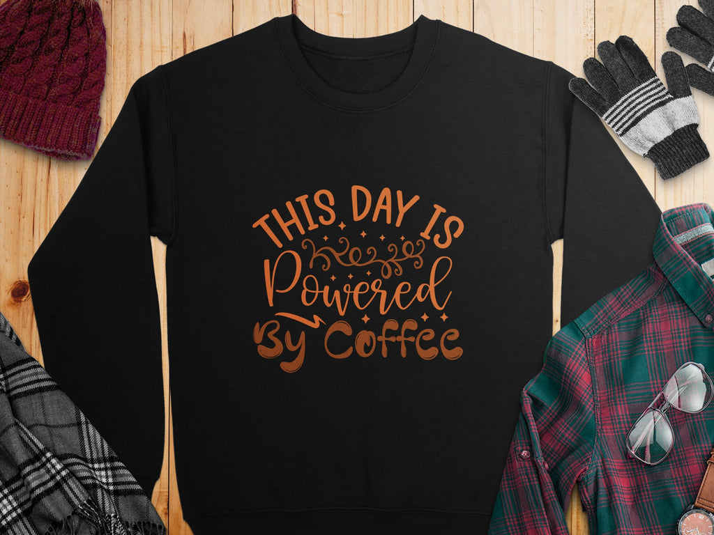 A black Garment Graphics sweatshirt with This Day is Powered by Coffee in bold orange font on wood, surrounded by a red knit hat, black gloves, a multi-colored plaid shirt, and gray and plaid fabric sections.
