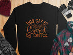 A black Garment Graphics sweatshirt with This Day is Powered by Coffee in bold orange font on wood, surrounded by a red knit hat, black gloves, a multi-colored plaid shirt, and gray and plaid fabric sections.