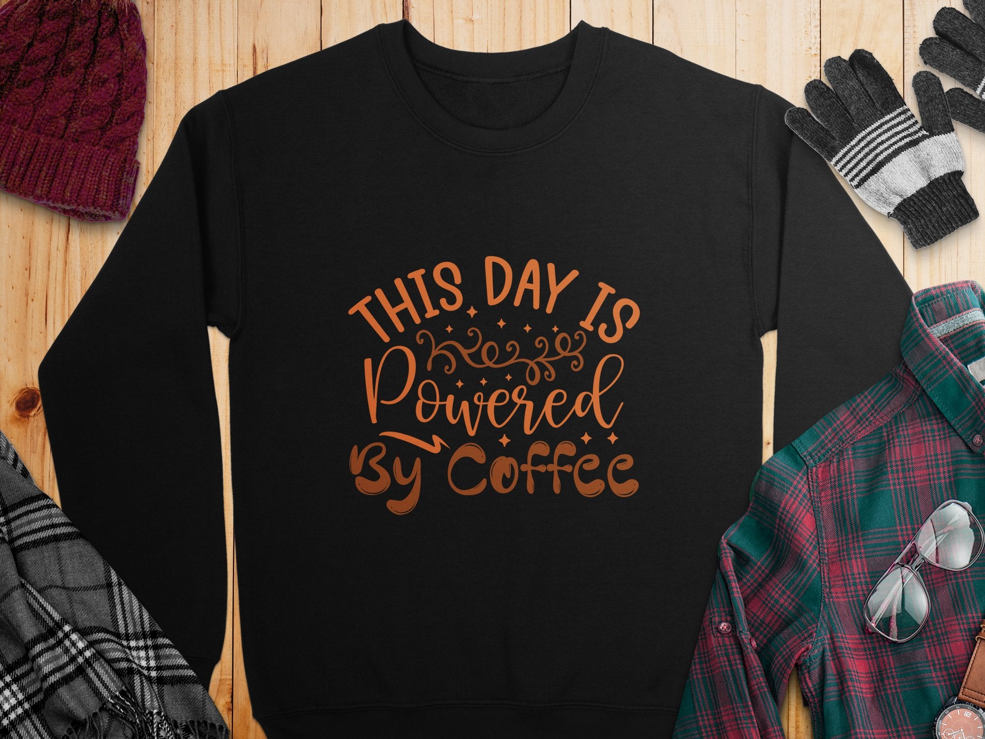 A black Garment Graphics sweatshirt with This Day is Powered by Coffee in bold orange font on wood, surrounded by a red knit hat, black gloves, a multi-colored plaid shirt, and gray and plaid fabric sections.