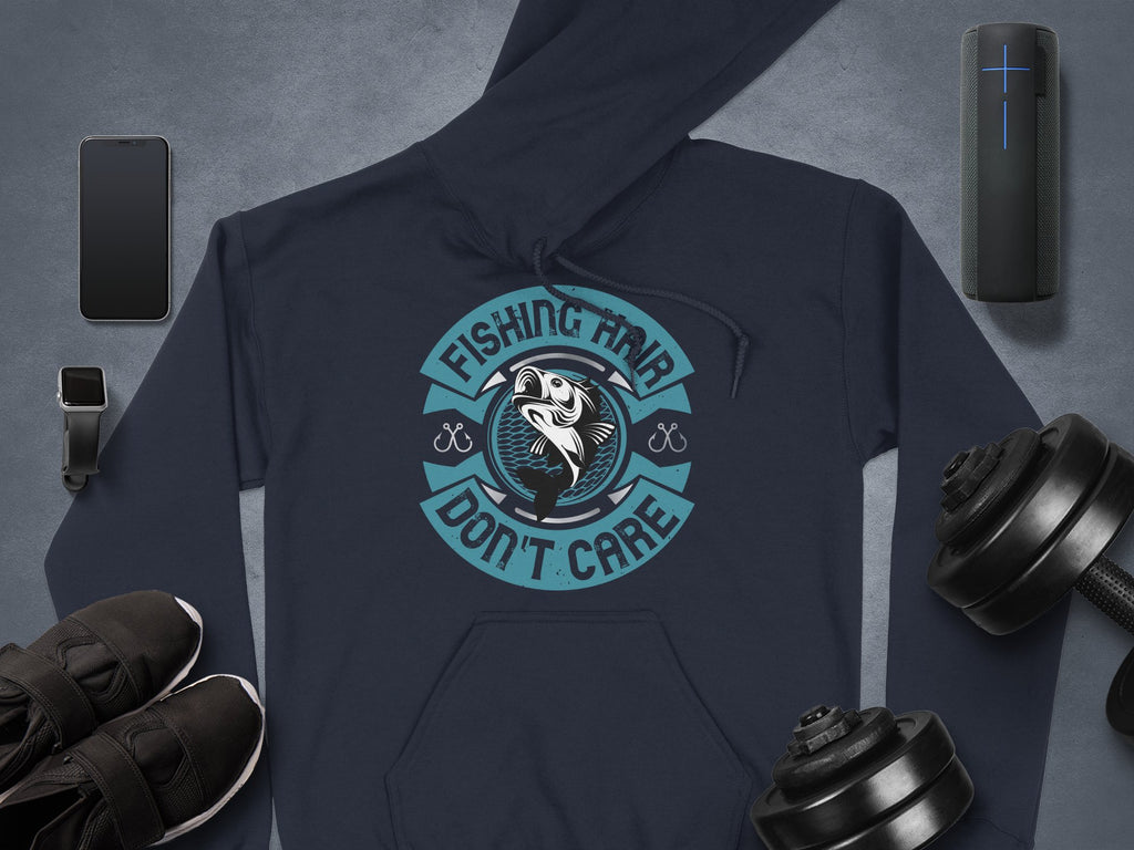 A dark Garment Graphics hoodie with a circular fish and crossed hooks logo and the text Fishing Hair Dont Care, perfect for anglers. Pictured on a gray surface are also a smartphone, smartwatch, speaker, sneakers, and adjustable dumbbells.