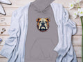 A bold statement piece from Garment Graphics, this gray hoodie featuring a cartoon bulldog with a spiked collar is on display on wood. Its stylishly paired with a light gray cardigan, white heels, a ring, bracelet, and delicate white flowers.