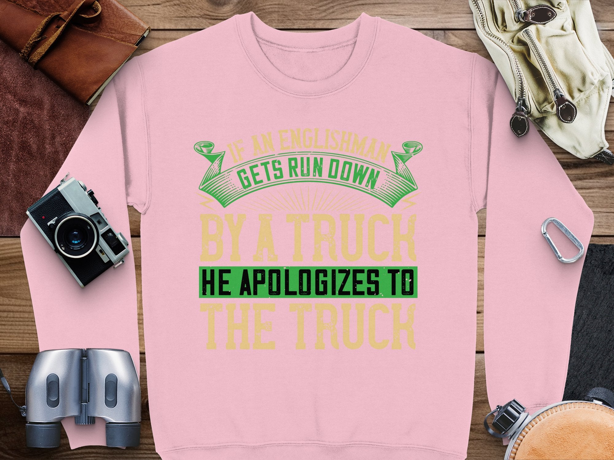 A pink unisex sweatshirt by Garment Graphics features a bold, humorous design: If an Englishman gets run down by a truck, he apologizes to the truck. Displayed on a wooden surface with a camera, binoculars, leather bag, notebook, and gloves.