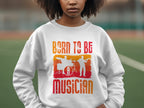 A person wears a Garment Graphics white sweatshirt with Born to Be Musician in colorful print, featuring drummer and musician silhouettes. Its perfect for music lovers and available in unisex sizing. They stand on an outdoor sports field.