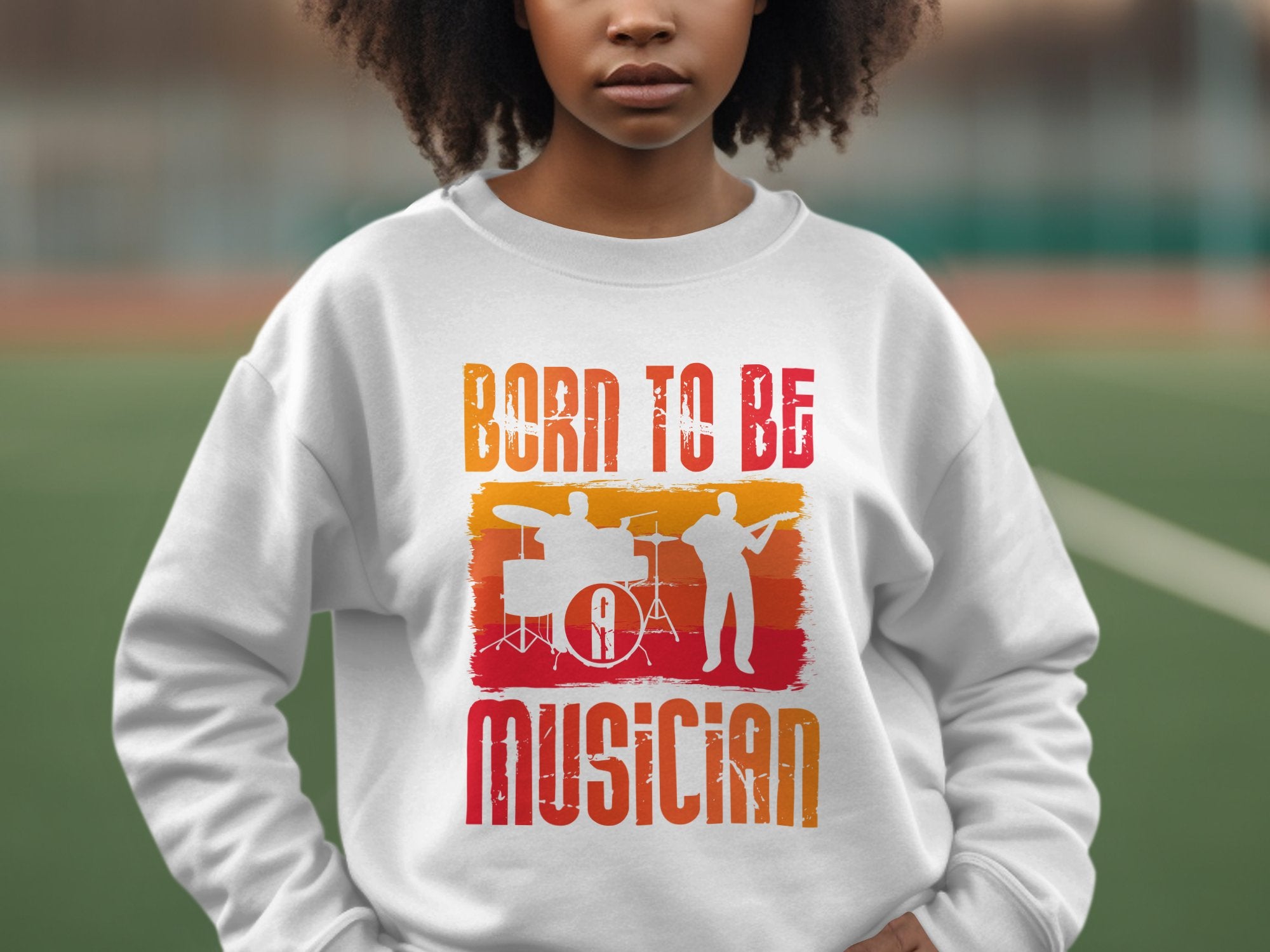 A person wears a Garment Graphics white sweatshirt with Born to Be Musician in colorful print, featuring drummer and musician silhouettes. Its perfect for music lovers and available in unisex sizing. They stand on an outdoor sports field.
