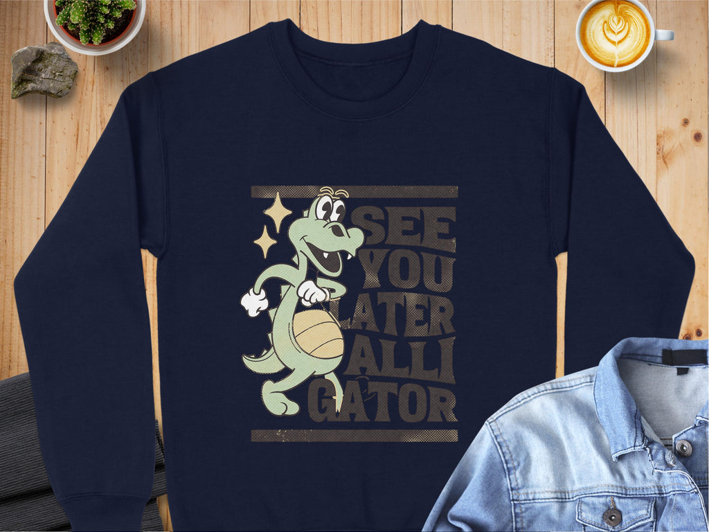 A Garment Graphics navy sweatshirt features a playful alligator holding a basketball, with See You Later Alligator on display. Its styled with a denim jacket, beside a potted plant and latte art on a wooden surface.