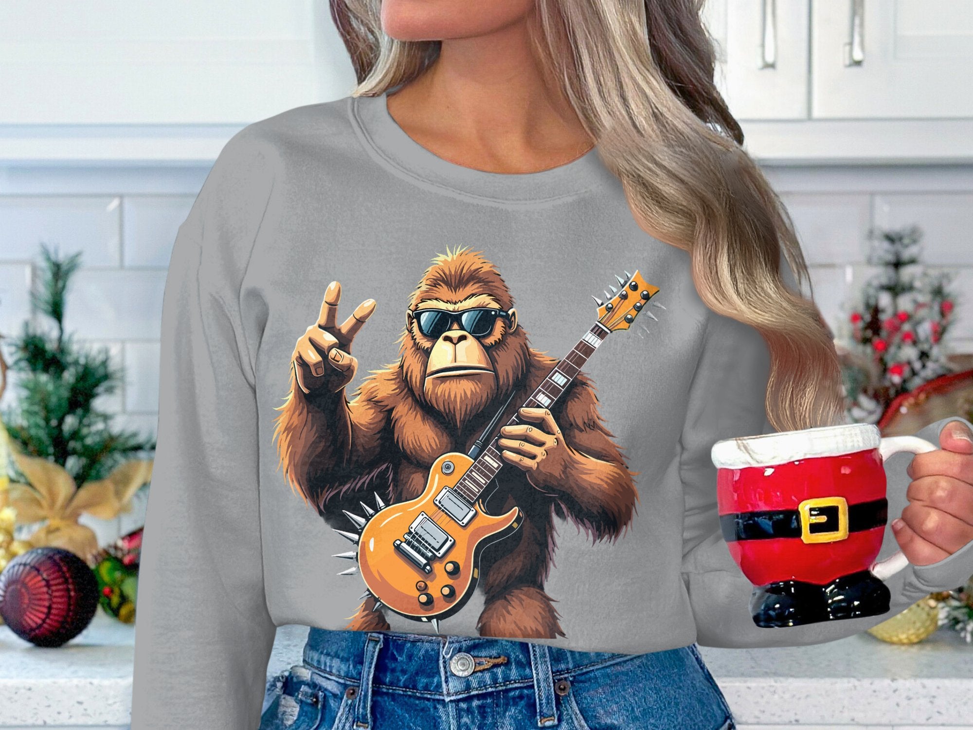 In a festive kitchen setting, someone dons a Garment Graphics sweatshirt with a gorilla playing an electric guitar, making a peace sign, and wearing sunglasses, while holding a Santa-themed mug. Holiday decorations fill the background.