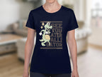 A person is wearing a Garment Graphics navy blue T-Shirt featuring a cartoon alligator holding a basketball and the funny text SEE YOU LATER ALLIGATOR, set against a blurred living room backdrop. The playful design is ideal for women.