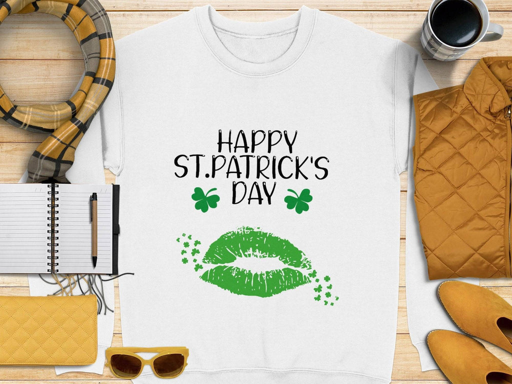 A Garment Graphics white t-shirt embodies the festive spirit with Happy St. Patricks Day in black, a green clover kiss, and two shamrocks, displayed on a wooden surface alongside a yellow jacket, scarf, purse, sunglasses, an open notebook with a pen, and a coffee cup.