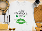 A Garment Graphics white t-shirt embodies the festive spirit with Happy St. Patricks Day in black, a green clover kiss, and two shamrocks, displayed on a wooden surface alongside a yellow jacket, scarf, purse, sunglasses, an open notebook with a pen, and a coffee cup.