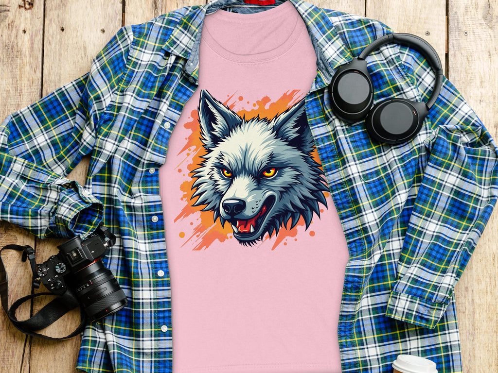 A 100% cotton pink T-shirt featuring a wolf print from Garment Graphics rests on wood, topped with an open blue plaid shirt; nearby are black headphones, a camera, and a lidded white paper cup, exuding daring vibes.