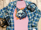 A 100% cotton pink T-shirt featuring a wolf print from Garment Graphics rests on wood, topped with an open blue plaid shirt; nearby are black headphones, a camera, and a lidded white paper cup, exuding daring vibes.
