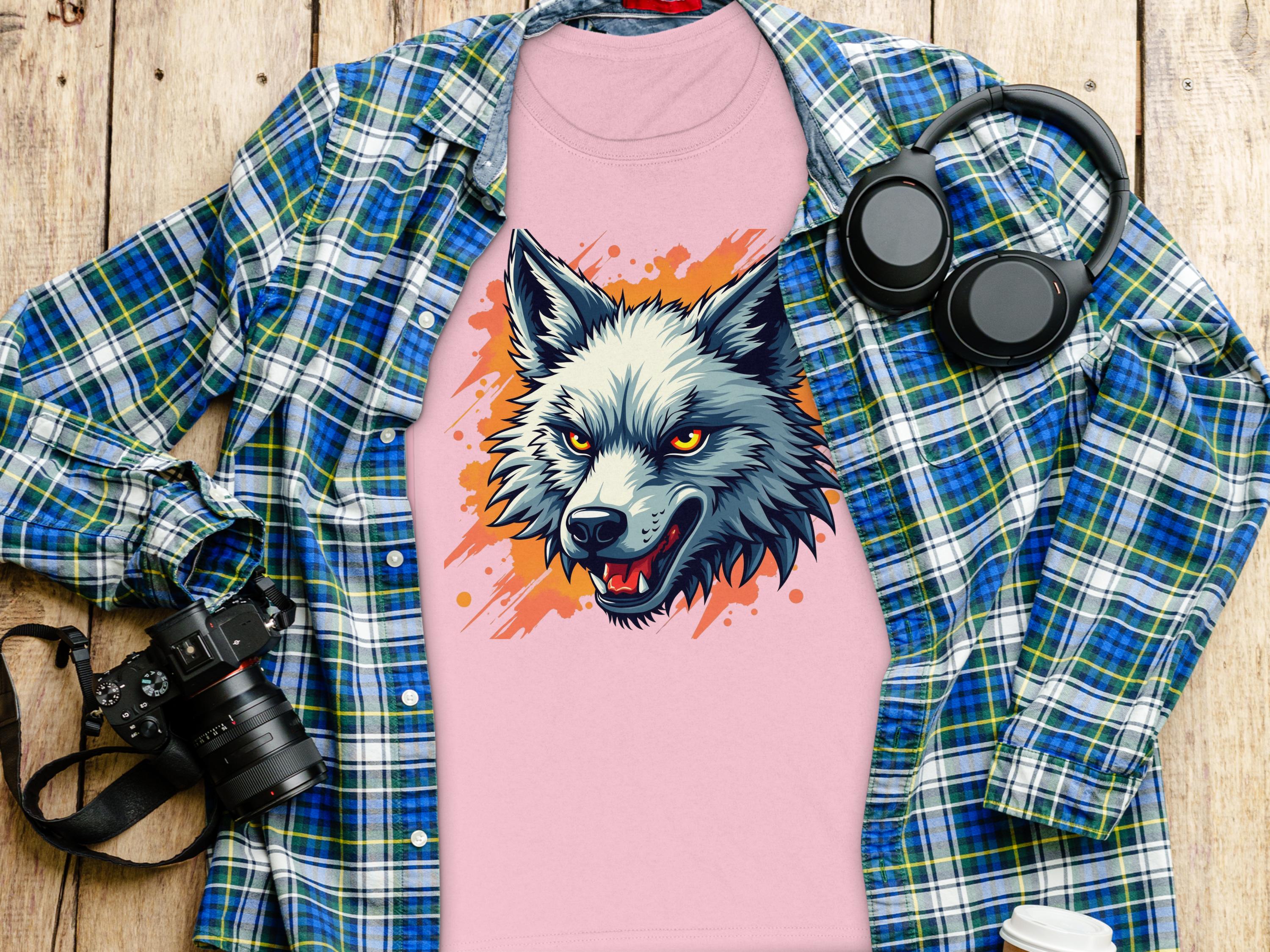 A 100% cotton pink T-shirt featuring a wolf print from Garment Graphics rests on wood, topped with an open blue plaid shirt; nearby are black headphones, a camera, and a lidded white paper cup, exuding daring vibes.