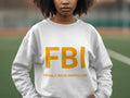 Someone wearing a Garment Graphics white unisex graphic sweatshirt stands outside. The sweatshirt displays large yellow FBI letters with smaller text Female Boob Inspector, against a blurred greenery and track backdrop.