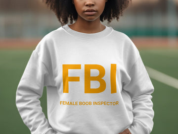 Someone wearing a Garment Graphics white unisex graphic sweatshirt stands outside. The sweatshirt displays large yellow FBI letters with smaller text Female Boob Inspector, against a blurred greenery and track backdrop.