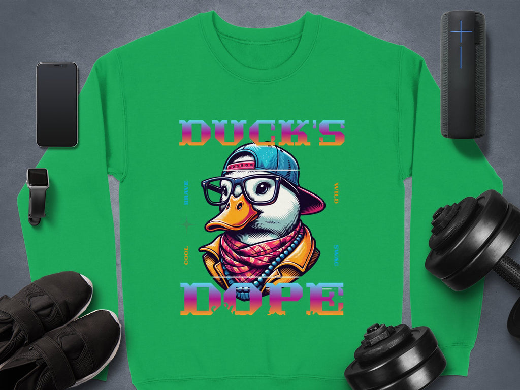 Garment Graphics green sweatshirt features a bold cartoon duck with sunglasses, a cap, and scarf. The Ducks Dope text enhances the design. Nearby essentials like a phone, smartwatch, speaker, shoes, and dumbbells lay on a sleek gray surface.