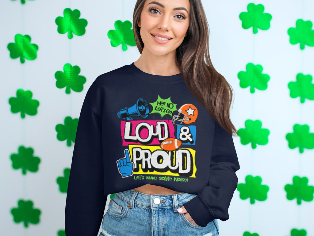Wearing a Garment Graphics Loud and Proud Sweatshirt in unisex sizing, adorned with vibrant megaphone and football graphics alongside LOUD & PROUD, a person with long brown hair and denim jeans stands against shamrock decorations, displaying their unique style.