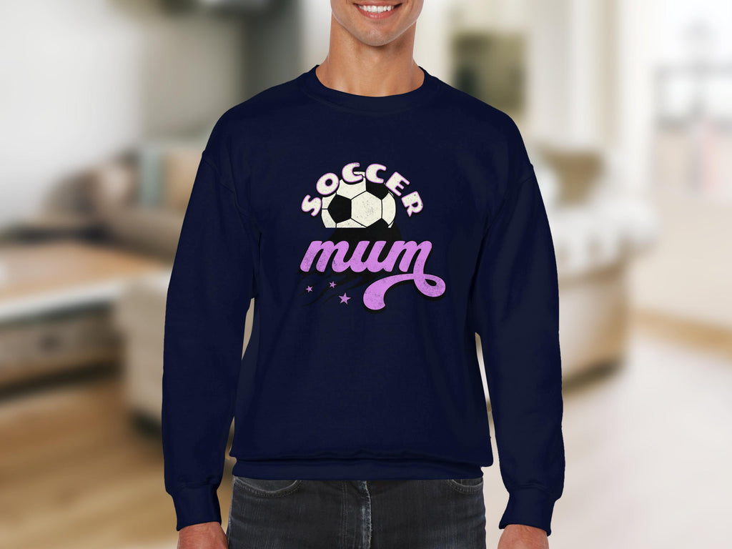 A person wears a Garment Graphics navy sweatshirt showcasing a vintage soccer ball design with Soccer Mum in white and pink, accented by decorative stars, set against a blurred living room background.