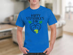 Wearing a Garment Graphics blue T-shirt with Happy St. Patricks Day and a heart and shamrock design radiates festive flair. The blurred indoor setting adds mystery to the cheerful celebration.