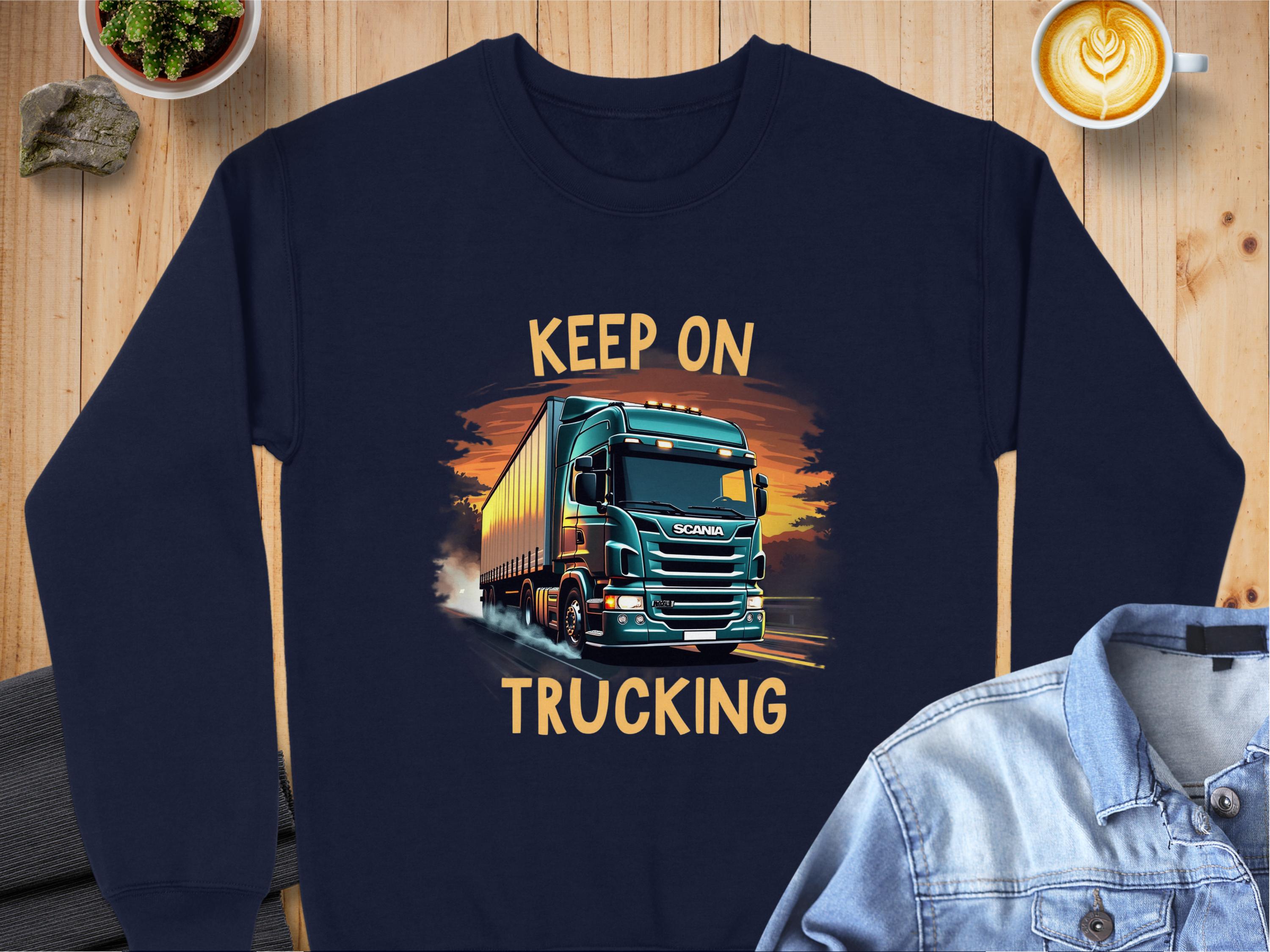 The Garment Graphics navy blue graphic sweatshirt features a large truck on a road with bold yellow Keep On Trucking text, ideal for truck fans. It pairs stylishly next to a denim jacket and latte on a wooden surface.
