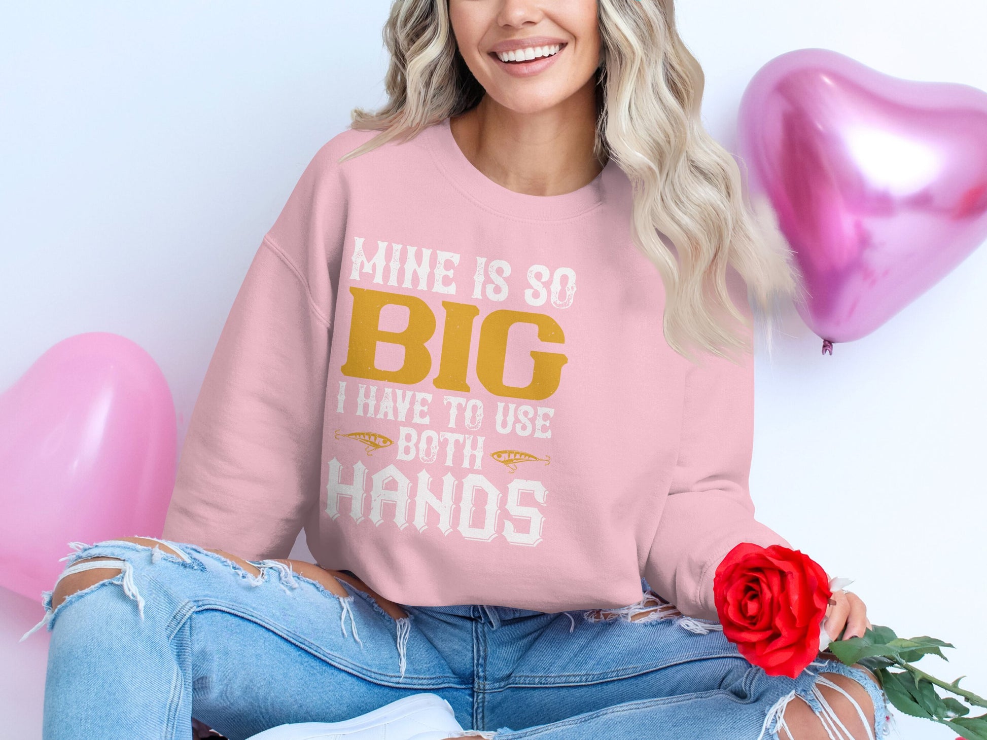 A woman in a distinctive pink Garment Graphics sweatshirt with the phrase Mine Is So Big I Have To Use Both Hands, sits cross-legged. Holding a red rose, shes surrounded by pink heart-shaped balloons and wears ripped jeans, smiling warmly in this playful scene.
