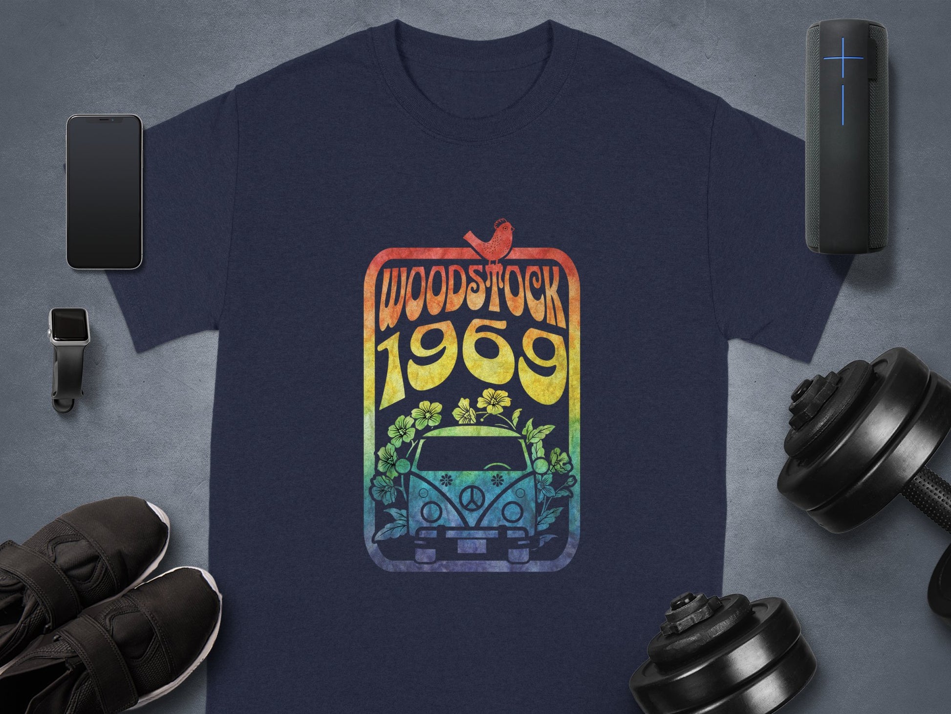 Garment Graphics presents a navy blue T-shirt with Woodstock 1969, a bird, and a retro van with flowers. Ideal for music lovers, it complements your smartphone, wireless speaker, smartwatch, sneakers, and weights for a harmonious lifestyle.