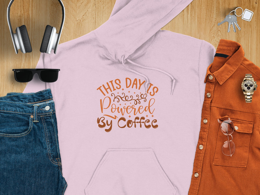 A flat lay features Garment Graphics’ light pink hoodie with This Day is Powered by Coffee text, ideal for coffee lovers. It’s styled with jeans, black sunglasses, large headphones, a rust button-up shirt and glasses, a watch, and keys on wood.